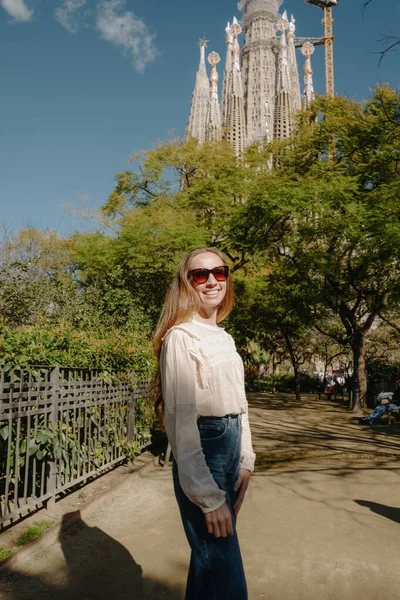 Parkta gülümseyen genç bir kadın boho bluz ve Sagrada Familia yakınlarında koyu renk kot pantolon giyiyor.