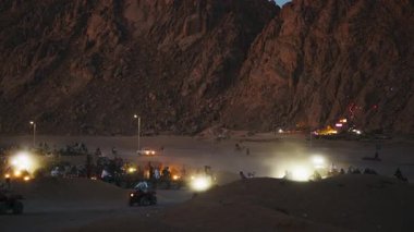 Bir grup turist ön plandaki tozlu yolda ATV 'ye biniyor ve ikinci grup arka planda park halinde. Gece görüş mesafesi kötü olduğu için araçların farları zaten açık..