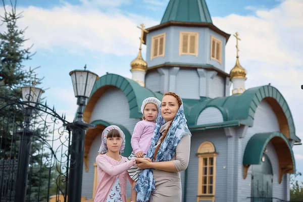 Russian Orthodox woman in a scarf stands with two daughters on the background of the Russian Orthodox Church.