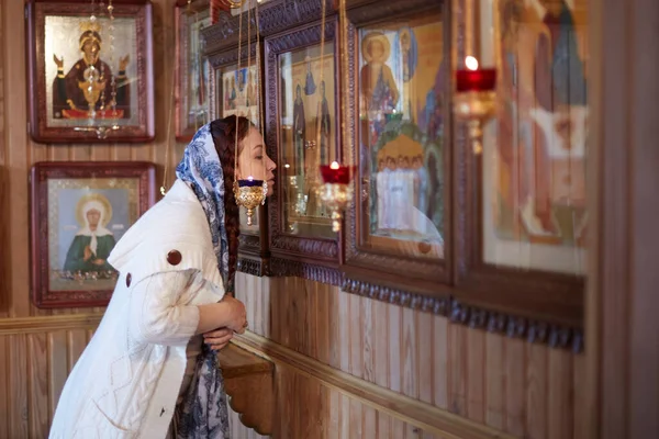 woman in the Russian Orthodox Church with red hair and a scarf on her head lights a candle and prays in front of the icon.