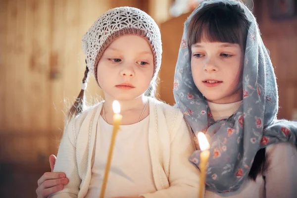 two Russian girls in a scarf on their heads stand in an Orthodox Church, lights a candle and prays in front of the icon.