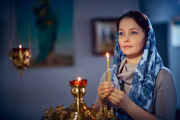 woman in the Russian Orthodox Church with red hair and a scarf on her head lights a candle and prays in front of the icon.