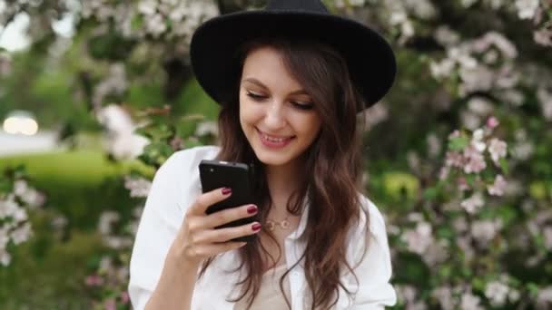 Mulher de pé no prado na árvore florescente, usando smartphone — Vídeo de Stock