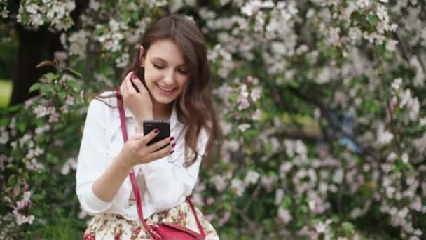 Mujer de pie en el prado en el árbol floreciente, utilizando el teléfono inteligente — Vídeo de stock