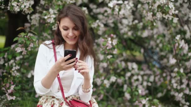 Жінка стоїть на лузі на квітучому дереві, використовуючи смартфон — стокове відео