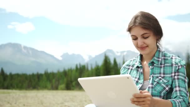 Feliz joven mujer morena caucásica con tableta en el parque en el soleado día de verano sentado en la hierba, de nuevo iluminado, sonriendo, mirando a la pantalla de la tableta. Conceptos modernos de estilo de vida y relajación . — Vídeos de Stock