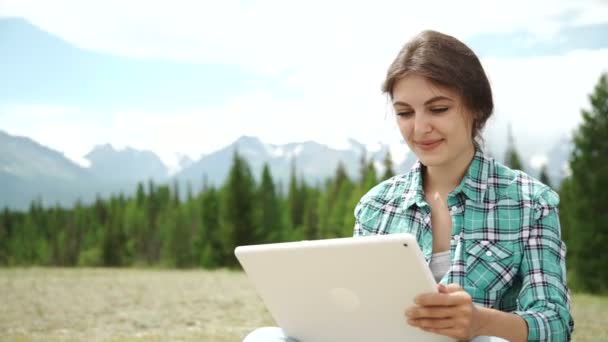 Glada unga kaukasiska brunett kvinna med tabletten i parken på solig sommardag sitter på gräset, tillbaka lit, leende, tittar på Tablet PC-skärmen. Modern livsstil och avkoppling begrepp. — Stockvideo