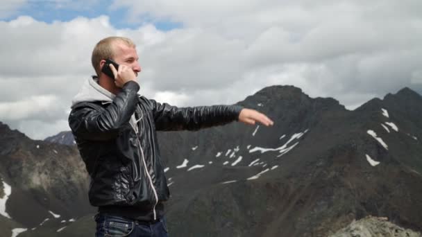 Porträt eines mutigen Mannes mit einem Rucksack, der auf einem Felsen sitzt, wegschaut und vor dem Hintergrund von Waldtal und Hügeln telefoniert. — Stockvideo