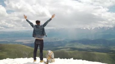 Sırt çantalı bir yürüyüşçü dağın tepesinde duruyor, elleri havada ve güneşin doğuşunun keyfini çıkarıyor.