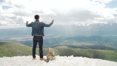 Sırt çantalı bir yürüyüşçü dağın tepesinde duruyor, elleri havada ve güneşin doğuşunun keyfini çıkarıyor..