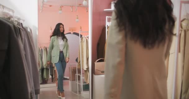 A Back View Shooting of a Brunette Woman Buyer Being in a Clothing Shop De pie frente al espejo y probándose una chaqueta. Hermosa alegre joven mujer eligiendo ropa — Vídeo de stock