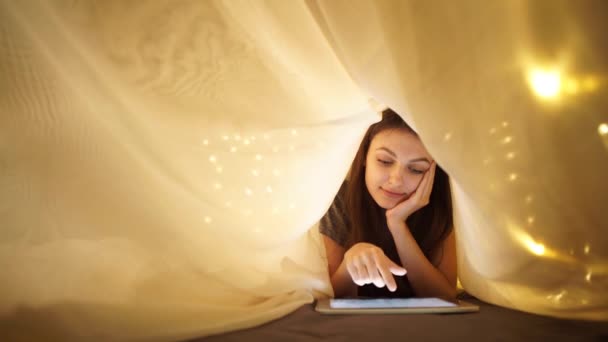 Девушка с цифровой планшет под одеялом, лежа на кровати дома — стоковое видео