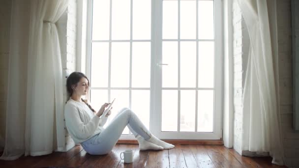 Молодая женщина дома сидит перед окном расслабляясь с помощью планшета — стоковое видео