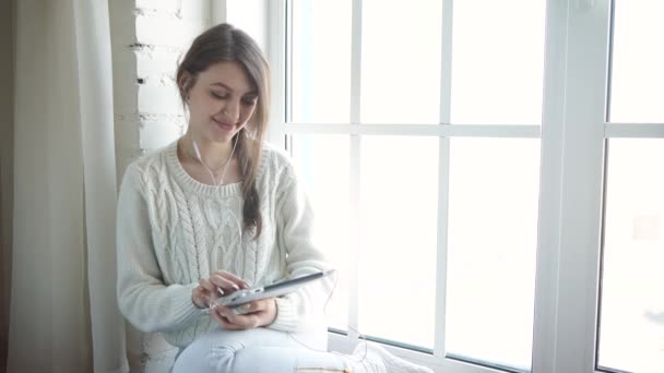 Молода жінка вдома сидить перед вікном, розслабляючись за допомогою планшета — стокове відео