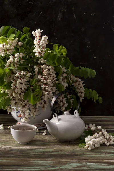 Still life with branches of a beautiful, delicate acacia tree with white clusters in a jug and aromatic tea in a white cup on a wooden table close-up