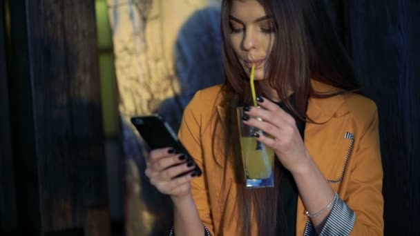 Morena bebiendo jugo, mirando por teléfono, sonriendo contra las cortinas del telón de fondo — Vídeos de Stock