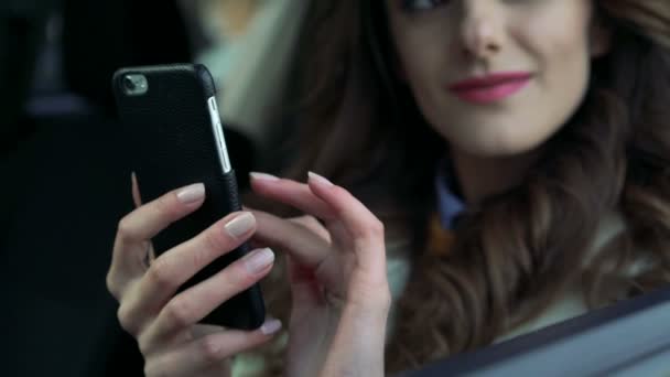 Девушка сидит в машине с телефоном, улыбается и смотрит в окно — стоковое видео