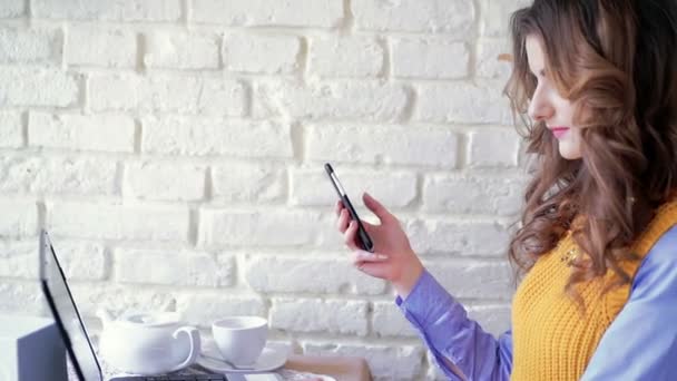 Mulher sentada no café, usando smartphone, laptop no backgroung da parede branca . — Vídeo de Stock