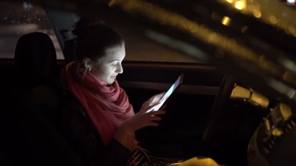 Chica bonita en una falda sentada en el coche, sonriendo y usando la tableta por la noche, 4K — Vídeos de Stock