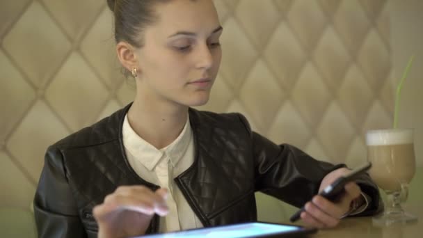 Chica sentada en la cafetería, usando una tableta, hablando por teléfono, sonriendo. 4K — Vídeos de Stock