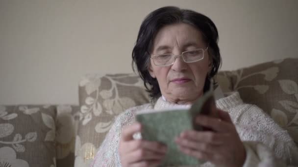 Старуха в очках читает молитвенник на диване — стоковое видео
