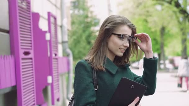 Chica fina sosteniendo una tableta, con gafas y sonriendo en el fondo de la calle 4k — Vídeo de stock