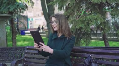 Bir tablet açık 4 k kullanan açık, bir bankta oturan kız gülümseyerek
