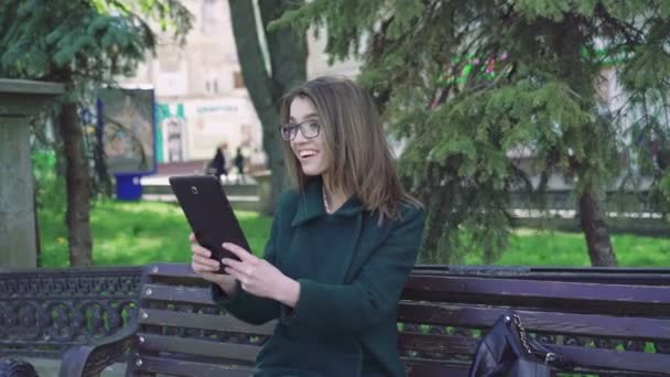 Χαμογελαστό κορίτσι συνεδρίαση σε έναν πάγκο εξωτερική, χρησιμοποιώντας μια ταμπλέτα εξωτερική 4k — Αρχείο Βίντεο