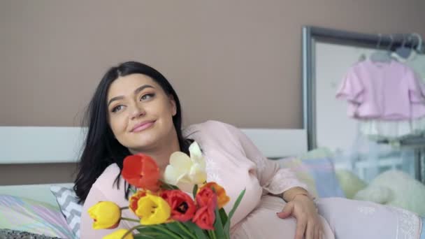 Милая беременная отдыхает на кровати, держа букет тюльпанов и улыбаясь. 4k — стоковое видео