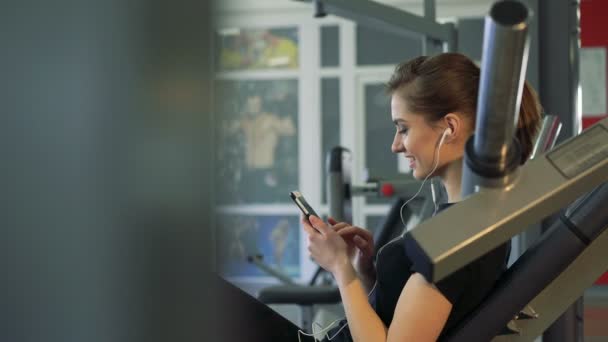 Primer plano de una chica en el gimnasio con un teléfono y un auricular — Vídeos de Stock