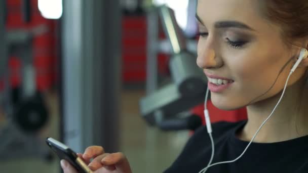 Joven mujer sonriente en el gimnasio escuchando música usando un teléfono móvil . — Vídeos de Stock