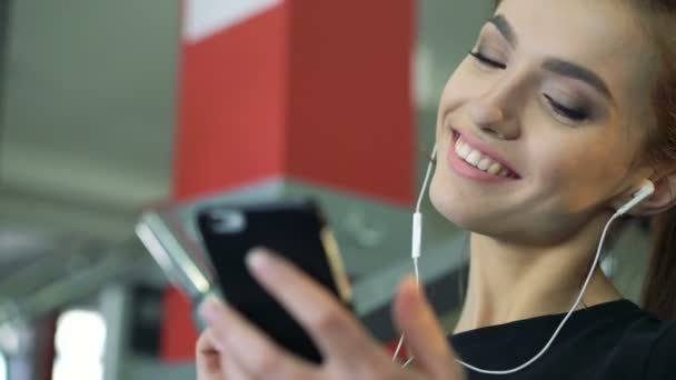 Joven mujer sonriente en el gimnasio escuchando música usando un teléfono móvil . — Vídeos de Stock