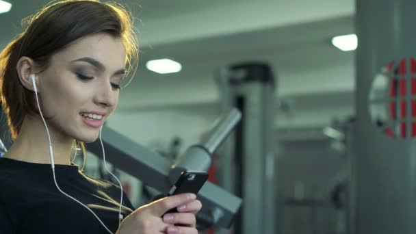 Primer plano de una chica en el gimnasio con un teléfono y un auricular — Vídeos de Stock