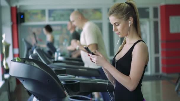 Красива блондинка на біговій доріжці використовує телефон і слухає музику. 4k — стокове відео
