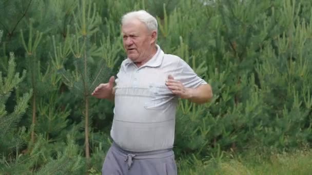 Oude man doet ochtend oefeningen rek handen vingers in park met coniferen — Stockvideo