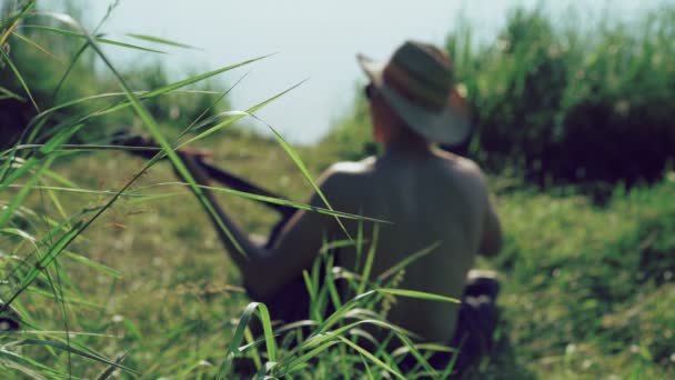 Mirada posterior de niño en gafas de sol y sombrero tocando la guitarra cerca del lago. 4k — Vídeo de stock