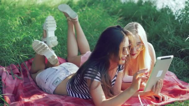 Брюнетка і блондинка лежать на ковдрах на траві і використовують планшет. 4k — стокове відео