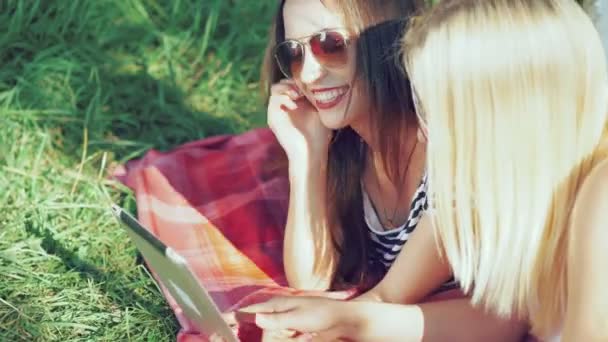 Две беззаботные девушки лежат на одеялах на траве и пользуются планшетом. 4k — стоковое видео