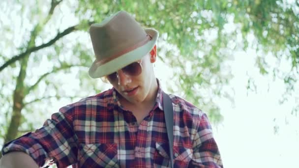 The young man in hat playing the guitar in sunny green park. 4k — Stock Video