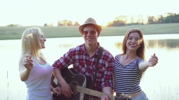 Ragazze che tengono le luci del bengala e ragazzo che suona la chitarra vicino al lago. 4k — Video Stock