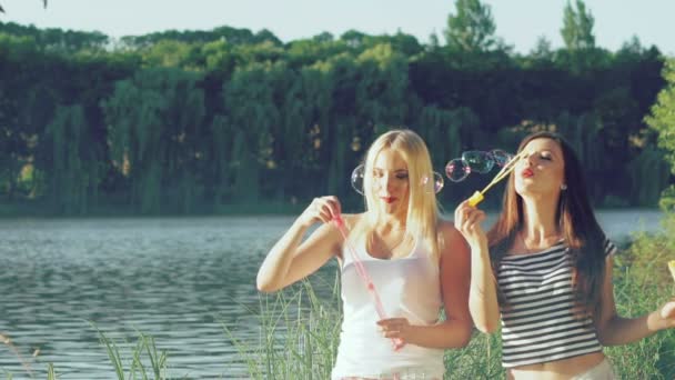 Chicas felices soplando burbujas de jabón de colores cerca del lago. Despacio. — Vídeos de Stock