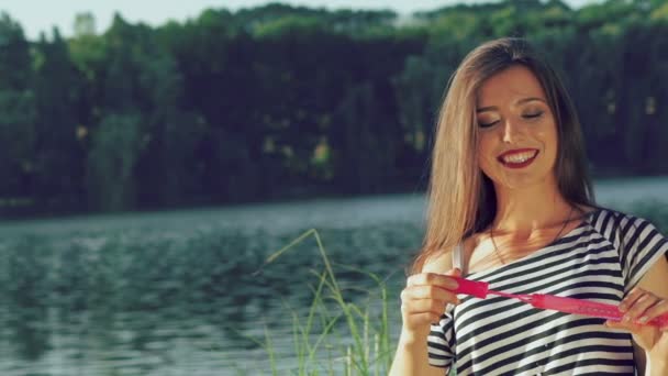 Joyeux brunette soufflant des bulles de savon colorées dans un parc d'été près d'un étang. Doucement. — Video