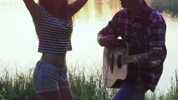 Chica joven bailando cuando niño tocando la guitarra cerca del lago. Despacio. — Vídeos de Stock