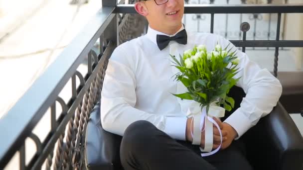 Молодой человек сидит со свадебным букетом на балконе — стоковое видео