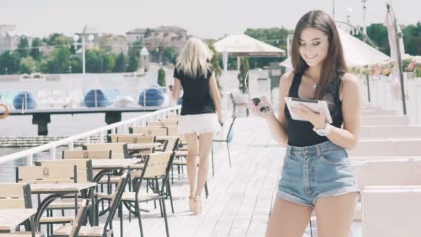 Chica en gafas de sol de pie en el muelle, utilizando la tableta y sonriendo. Despacio. — Vídeos de Stock