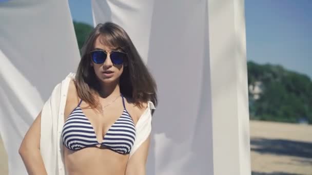 Sexy chica en traje de baño y gafas de sol posando cerca del mar en gazebo blanco. 4K — Vídeo de stock