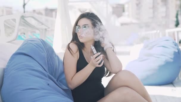 Chica en negro fumar cigarrillo electrónico, descansando en el salón y mirando a un lado en 4K — Vídeos de Stock