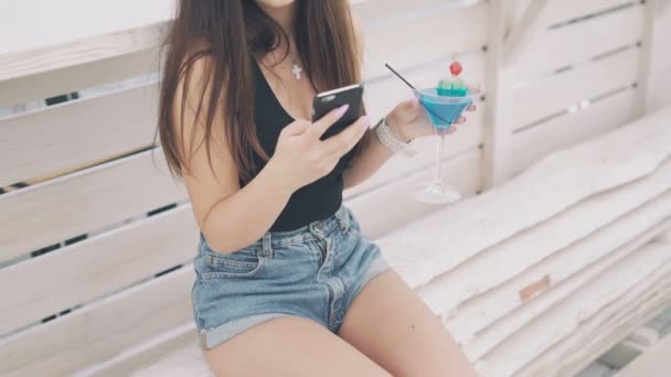 Молодая девушка с телефоном с бокалом коктейля в руке на скамейке на пляже в 4K — стоковое видео