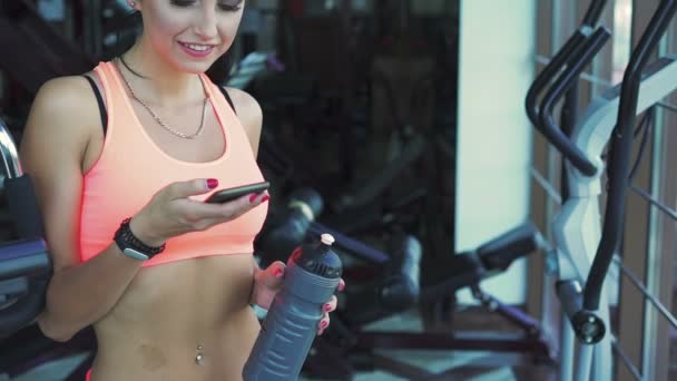 Happy woman holds bottle of water and using phone in a gym — Stock Video