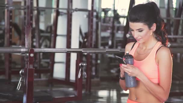 Junges Mädchen hält Wasserflasche in der Hand und benutzt Telefon in einem Fitnessstudio. langsam — Stockvideo